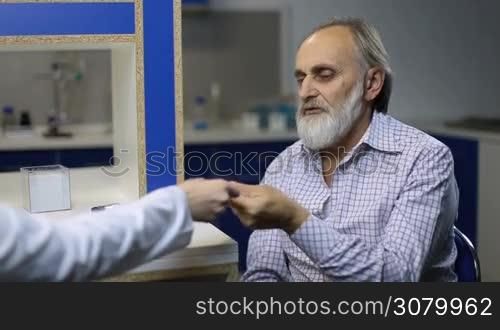 Senior ill patient with beard putting electronic thermometer under armpit while checking body temperature in doctor&acute;s office. Old sick man explraining symptoms to doctor during a visit physician at clinic