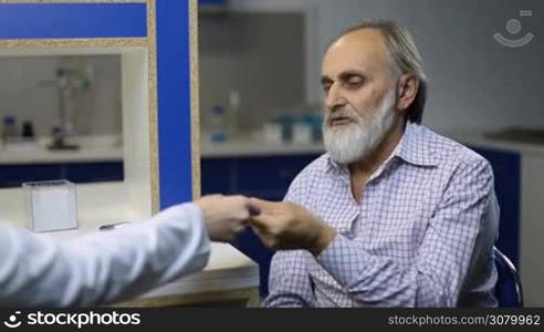 Senior ill patient with beard putting electronic thermometer under armpit while checking body temperature in doctor&acute;s office. Old sick man explraining symptoms to doctor during a visit physician at clinic