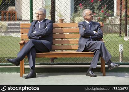 Senior male business executives looking away with arms crossed while sitting on bench in a tennis court
