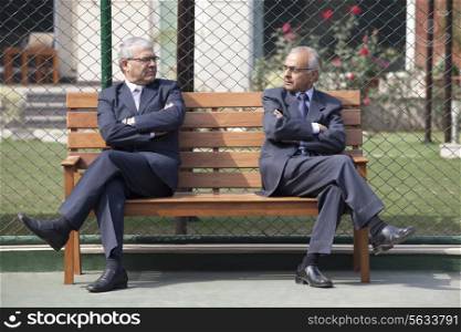 Senior male business executives staring at each other while sitting arms crossed on bench in tennis court