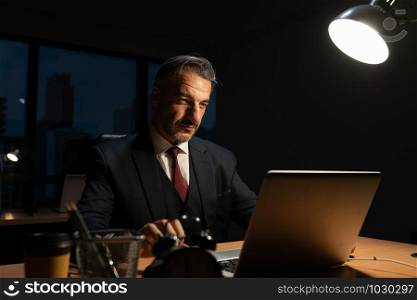 Senior male businessman in executive positions sitting working at office desk with laptop, While the works overtime in the office at night