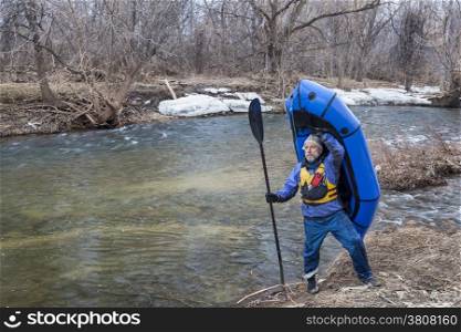senior male carrying a packraft (one-person light raft used for expedition or adventure racing) on the shore of Cache la Poudre River in Fort Collins, Colorado, winter or early spring scenery