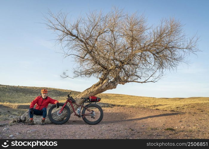 senior male cyclist with a fat mountain bike is resting at a lone tree in northern Colorado prairie, early spring scenery in Soapstone Prairie Natural Area near Fort Collins
