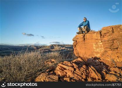 senior male hiker is sitting on a top of sandstone rock formation and watching sunset over Red Mountain Open Space in northernn Colorado