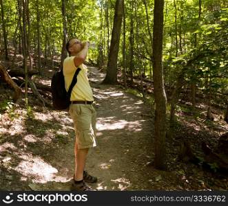Senior male hiker overlooking the path through forest and drinking from water bottle