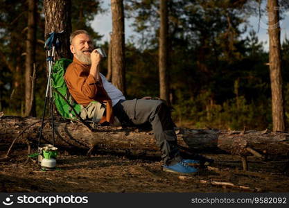 Senior male hiker taking coffee break during active walk adventure outdoors in pine forest. Retired man drinking warm beverage after lunch. Weekends on nature during retirement time. Senior male hiker taking coffee break during active walk adventure outdoors