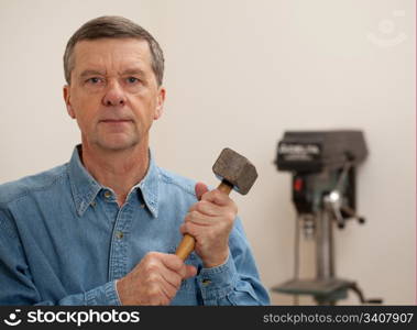 Senior male in a home workshop facing the camera and holding a large lump hammer