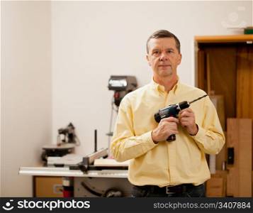 Senior male in a home workshop facing the camera and holding a power drill