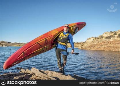 senior male is carrying a colorful river kayak on a rocky lake shore Horsetooth Reservoir) - recreation concept