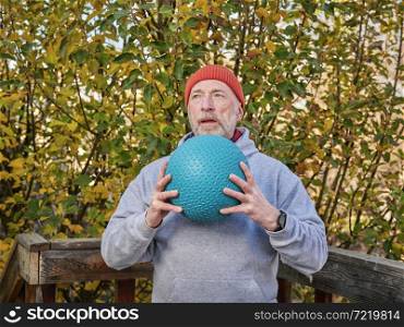 senior male (on late 60s) is exercising with a heavy slam ball in his backyard, chilly afternoon in fall scenery