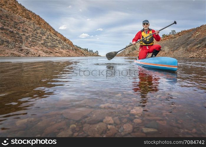 senior male paddler in drysuit is enjoying stand up paddling on lake in Colorado, winter scenery with some ice