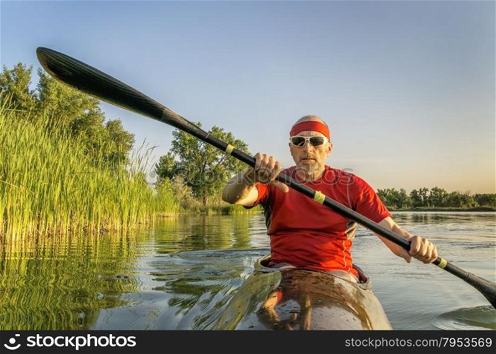 Senior male paddler is paddling a racing sea kayak on a lake along shore covered by reed