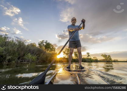 senior male paddler is paddling stand up paddleboard at sunset on a lake in Colorado