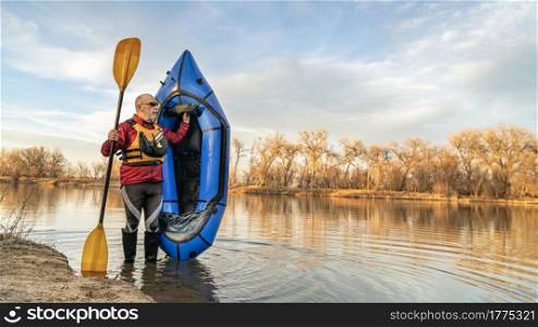 senior male paddler is standing with an inflatable packraft and paddle on a lake shore in early spring spring in northern Colorado