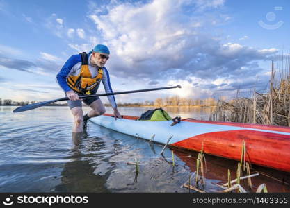 senior male paddler with his stand up paddleboard on a shore of calm lake, solo paddling as fitness and training with social distancing