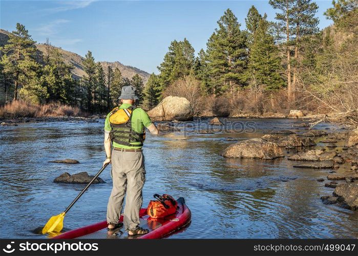 senior male paddling inflatable stand up paddleboard through a rock garden on a mountain river - Poudre RIver in Colorado in spring scenery with low water flow