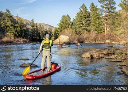 senior male paddling inflatable stand up paddleboard through rock garden on a mountain river - Poudre RIver in Colorado in spring scenery with low water flow
