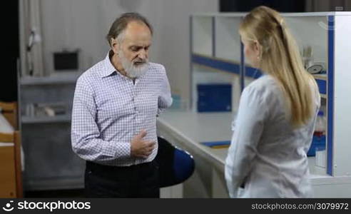 Senior male patient with beard suffering with low backache explaining to attentive female physiotherapist the cause of his pain during medical exam. Elderly man visiting a doctor office due to suffer terrible back pain.