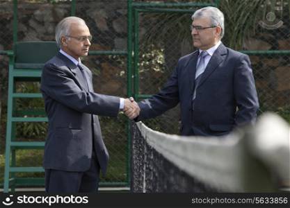 Senior male professionals staring at each other while shaking hands over tennis net in court