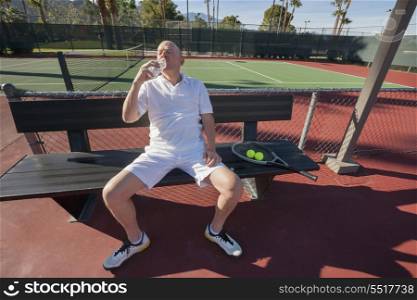 Senior male tennis player drinking water while relaxing on court