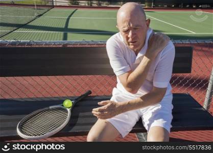 Senior male tennis player with shoulder pain sitting on bench at court