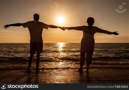 Senior man and woman couple holding hands arms wide at sunset or sunrise on a deserted tropical beach