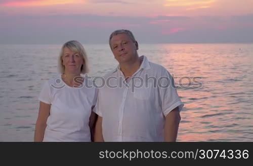 Senior man and woman embracing and looking to the camera with smile, evening sky and quiet sea in background
