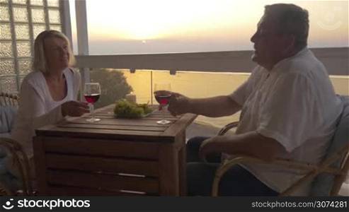 Senior man and woman enjoying time face to face. They having red wine relaxing in chairs on the outdoor balcony at sunset. Romantic evening