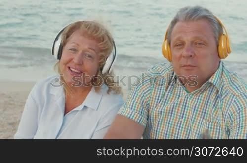 Senior man and woman listening to music in wireless headphones by the sea. They enjoying songs as dancing with favorite rhythms. Relaxing outdoor
