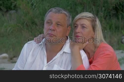 Senior man and woman outdoor. She embracing him over the shoulders and they staring somewhere far with dreamy and thoughtful look