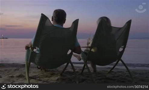 Senior man and woman sitting in chaise longues at the seaside in the evening. They clanging glasses with cocktails and relaxing looking at the sea