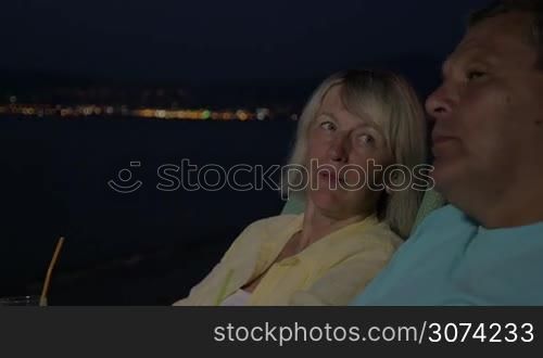 Senior man and woman spending summer evening at the seaside at night. They drinking cocktails, having lively conversation and looking at something distant