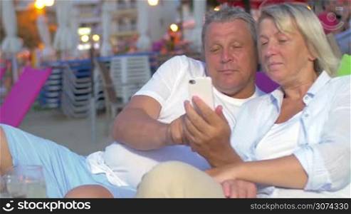 Senior man and woman using mobile phone while relaxing on the beach in the evening. Outdoor cafe in background
