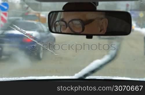 Senior man driving a car in the city in winter. Melting and dirty snow on the road. Driver in glasses reflecting in rear mirror