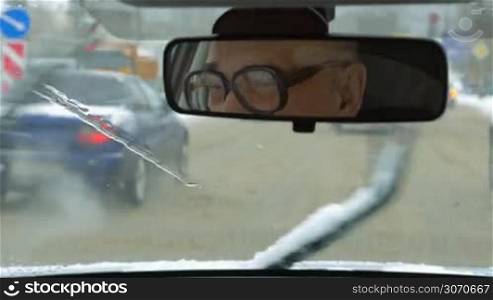Senior man driving a car in the city in winter. Melting and dirty snow on the road. Driver in glasses reflecting in rear mirror