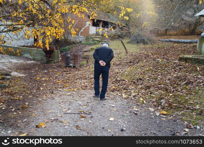 Senior man farmer at the farm walking on the dirt road in autumn day going to the piggery to feed the animals back view old pensioner