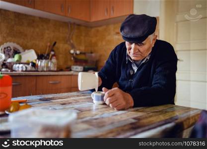 Senior man grandfather old pensioner farmer wearing black sweater and hat having a cup of coffee or tea by the table at home sitting alone pouring coffee from the pot
