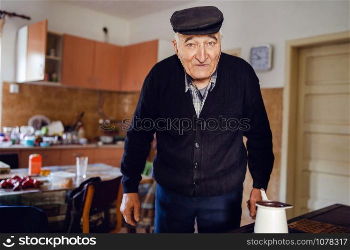 Senior man grandfather old pensioner farmer wearing black sweater and hat having a cup of coffee or tea cooking in the pot at home waiting to boil