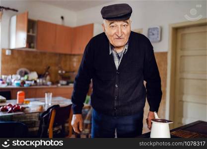 Senior man grandfather old pensioner farmer wearing black sweater and hat having a cup of coffee or tea cooking in the pot at home waiting to boil