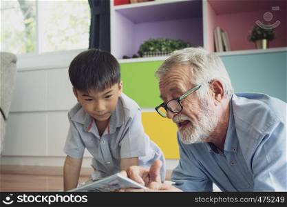 senior man happiness and grandson are sitting on the floor and playing games and reading book at living room together