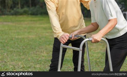 Senior man helping beloved wife walking with walker frame while doing rehab physical in the park.