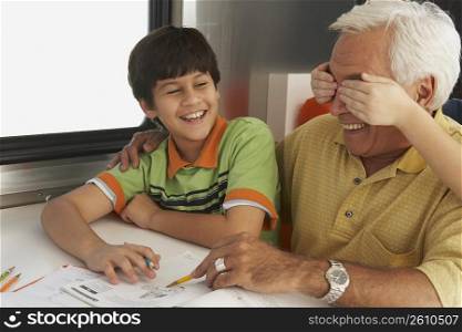 Senior man helping his grandson in his homework and a child&acute;s hands covering his eyes from behind