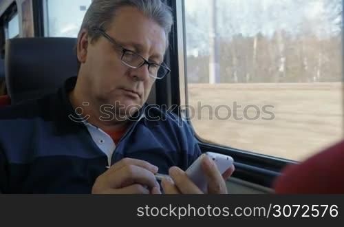 Senior man in glasses using pen and smartphone to browse the internet while traveling by train. Passing the time during the trip