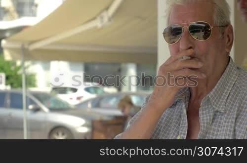Senior man in sunglasses holding cigarette in trembling hands and smoking sitting in the city street