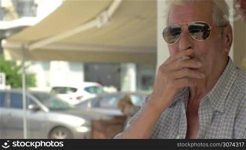 Senior man in sunglasses holding cigarette in trembling hands and smoking sitting in the city street