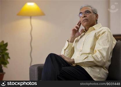 Senior man looking elsewhere with hand on cheek while sitting alone at home 