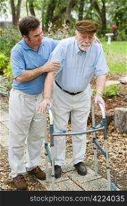 Senior man looks sad as he struggles to walk using a walker. His adult son is helping him.