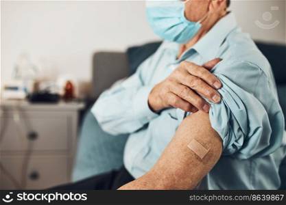 Senior man patient holding shirt s≤eve up with a plaster in place of injection of vacci≠. Covid-19 or coronavirus vaccination. Man wearing face mask
