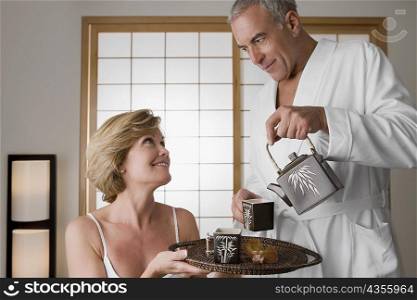 Senior man pouring tea from a tea kettle into the cup with a mature woman holding a tray