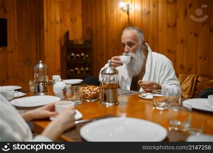 Senior man relaxes at dining table and flirting with young woman during rest in sauna. Senior man rest with friends in sauna